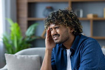 A young man with curly hair experiencing a headache, sitting indoors and holding his head in pain.
