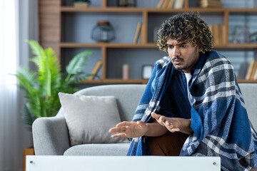 A man wrapped in a blanket sits indoors, possibly feeling cold, with his hands up in front of him.