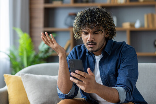 A confused man looks at his phone while sitting on a couch. He appears to be frustrated or perplexed.