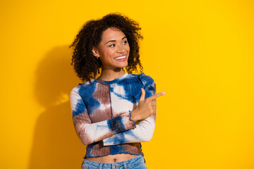 Young woman with curly black hair pointing on vibrant yellow background, wearing stylish casual...