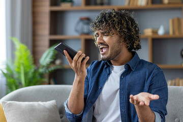 A man, indoors, expresses frustration while using his phone. He's on a call, gesturing with his...