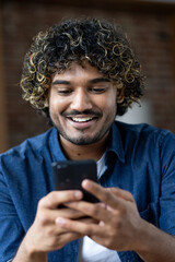 A smiling man of Indian descent is engrossed in his phone, likely texting or using an app indoors.