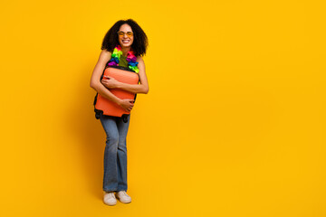 Smiling young woman holding an orange suitcase wearing vibrant lei and sunglasses, standing against...