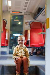 Young boy sits calmly on a step inside a modern train. Bright red seats and corridor behind create...
