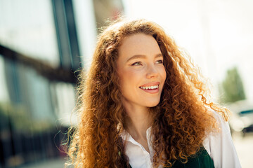 Elegant young woman with curly red hair smiling confidently outdoors in urban city on a sunny day