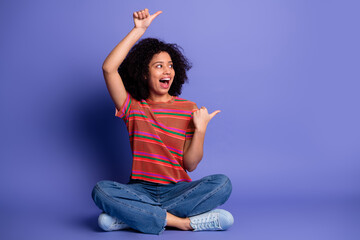 Happy young woman sitting cross-legged on a purple background pointing with enthusiasm and smiling...