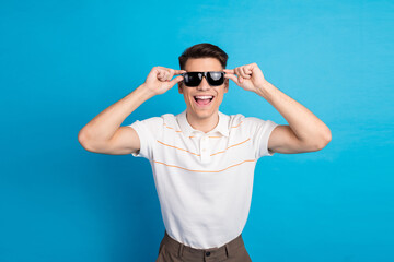 Young cheerful man in casual polo smiling and touching sunglasses, standing against a vibrant blue...