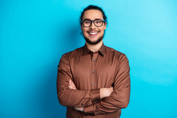 Confident young man in brown shirt poses with arms crossed against a turquoise background exuding...