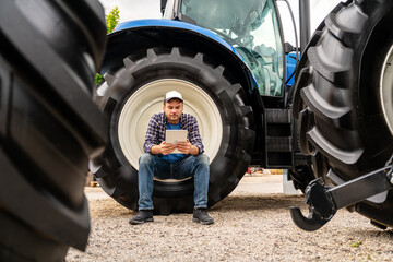 Male farmer using digital tablet while sitting on tractor wheel at agricultural dealership © Barillo Images