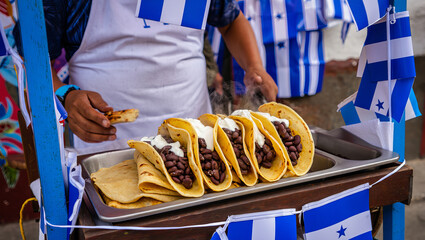 A vendor prepares traditional Honduran tacos filled with beans and cheese on a bustling street during a festive celebration. Colorful decorations enhance the lively atmosphere.