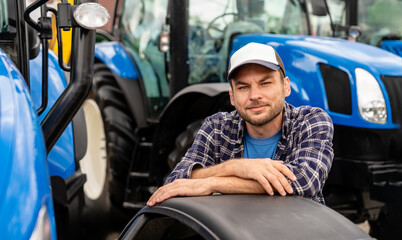 Portrait of young adult farmer in cap with tractor on background.