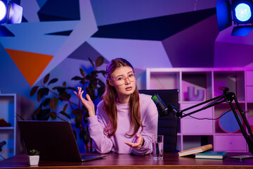 Young female blogger recording a live podcast at her desk with a microphone, laptop, and colorful...