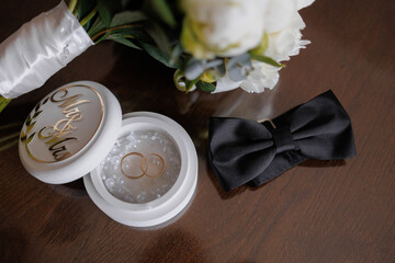 Beautiful wedding rings placed near a bouquet and a black bow tie on a wooden surface
