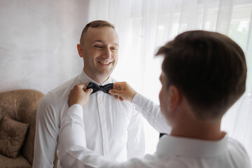 Groom gets ready for wedding ceremony while adjusting bow tie in a well-lit room