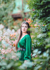 Elegant young woman in a green dress with a black belt posing in a lush garden filled with blooming...