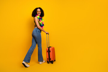 Happy young woman in casual clothing carrying an orange suitcase against a vibrant yellow...