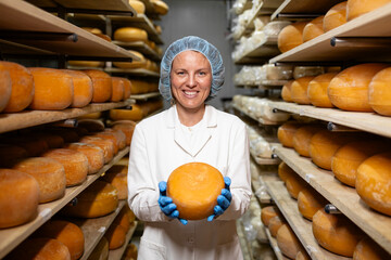 Portrait of female cheese maker holding cheese wheels. In background shelves full with dairy...