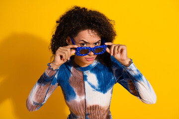 Stylish young woman holding funky blue sunglasses posing against a vibrant yellow background in a...
