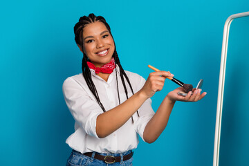 Young woman with braided hair smiling and applying makeup with a brush against a bright blue...