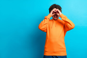 Cheerful boy in orange hoodie playfully looking through improvised binoculars against vibrant blue...