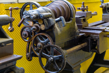 old screw-cutting lathe carriage with visible gears and handwheels. Industrial machine against a yellow metal background in a workshop.