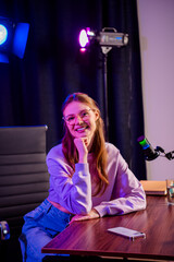 Charming young woman with glasses in a casual sweatshirt at a podcast setup under colorful neon...