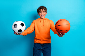 Cheerful young boy displaying a soccer ball and basketball in his hands on a vibrant blue background