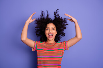 Joyful young woman with raised hands and curly hair against vivid purple background celebrating...