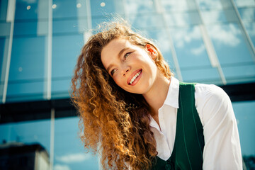 Confident young businesswoman with curly hair smiling cheerfully outdoors on a sunny day in front...