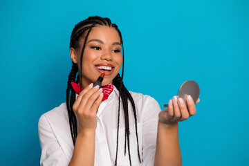 Smiling young woman applying lipstick while holding a compact mirror, standing against a vibrant...