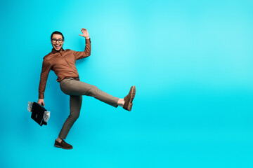 Dynamic young businessman jumping joyfully holding a black briefcase full of money on a bright teal...