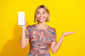 Cheerful blonde woman showcasing a smartphone against a vibrant yellow background in a stylish...