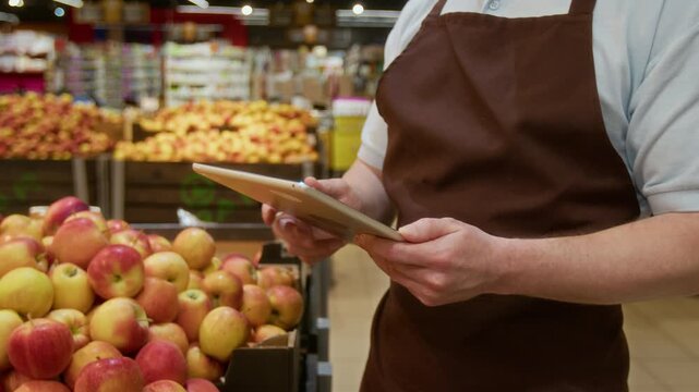 Midsection view of unknown male grocery worker wearing apron over T-shirt using digital tablet standing next to cardboard boxes filled with fresh apples during workday