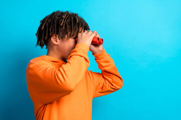 Young boy wearing orange hoodie looking through red binoculars against a bright blue background