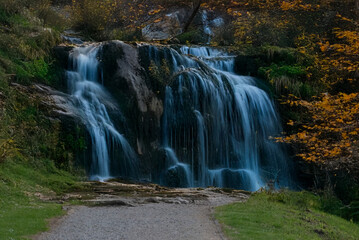 Fototapeta premium A multi-tiered waterfall cascades over mossy rocks surrounded by autumn foliage, ending at a gravel trail in a quiet, forested landscape.