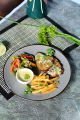 A delicious Grilled Fish With Sauteed Vegetables with French fries, salad, and a dipping sauce, presented on a dark plate with a textured background.