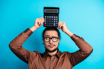 Young man holding calculator on head against turquoise background expressing thoughtful behavior in...