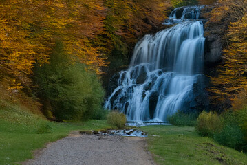 A majestic multi-tiered waterfall plunges through a forest in vibrant autumn colors, with a gravel path leading toward the cascading water in a serene setting.
