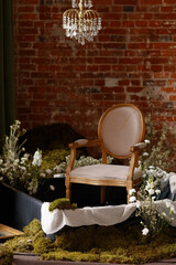 Classic wooden chair with beige fabric stands on moss and flowers under chandelier in rustic room...