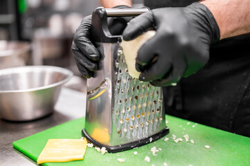 Close-up of gloved hands grating cheese on a metal box grater over a green cutting board in a...