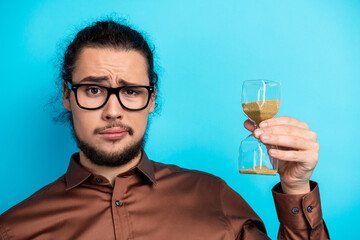 Young man with glasses holds an hourglass showing time's passing, expressing concern on a teal...