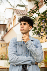 Denim fashion and relaxed vibes in garden setting. Young man in a denim jacket enjoys a cup of coffee in an urban garden, surrounded by plants and flowers, in a peaceful outdoor moment.