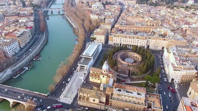 Stunning golden hour aerial footage showcasing the circular Mausoleum of Augustus beside modern Ara Pacis Museum along the Tiber River, framed by bridges and streets.