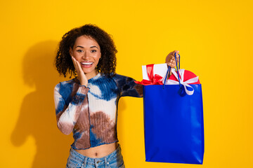 Happy young woman with gifts in a blue bag on a yellow background, exuding charm, surprise, and...