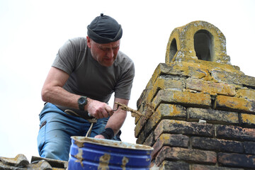 Experienced plasterer on roof of old house uses trowel and mortar to repair chimney. Sky in background. Handicraft.