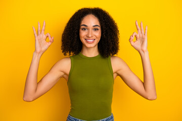 Smiling young woman in green top making okay gestures against bright yellow background showing...