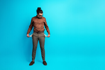 Young man in brown formal shirt on blue background displaying empty pockets, expressing financial...