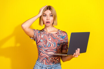 Blonde young woman in printed top holding laptop, surprised expression, standing against a vibrant...