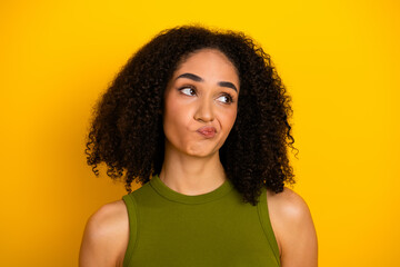 Young stylish woman with curly hair in green top against yellow background, expressing thoughtful...