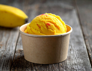 Yellow ice cream scoop in a paper cup, with a banana in the background, on a wooden surface.